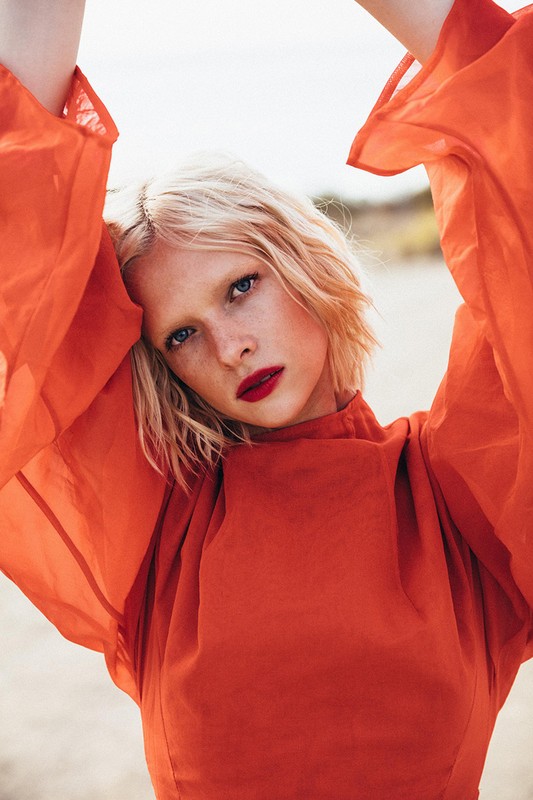 Close-up portrait of a woman with short blonde hair and bright red lipstick wearing an orange dress with voluminous sleeves against an outdoor background.