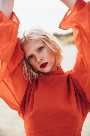Close-up portrait of a woman with short blonde hair and bright red lipstick wearing an orange dress with voluminous sleeves against an outdoor background.
