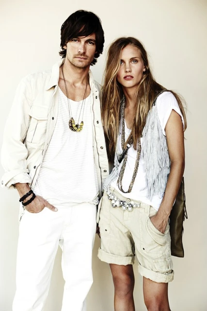 Two fashion models pose in a bright studio wearing casual white tops, shorts, jackets, and beaded necklaces against a neutral backdrop.
