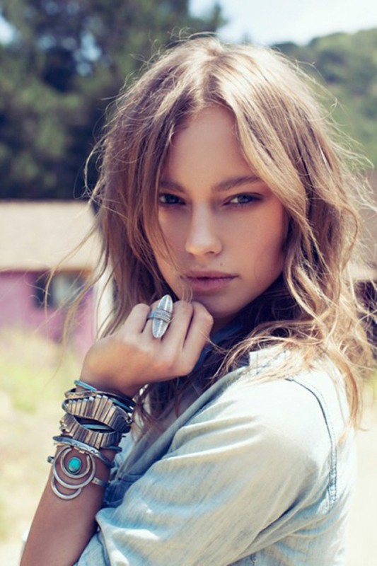 Close-up portrait of a woman with layered silver bracelets and a large statement ring, outdoors in daylight.