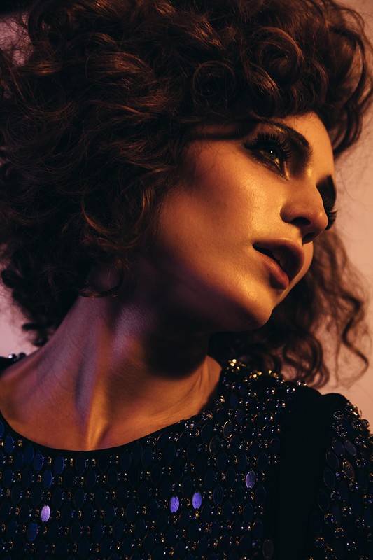 Portrait of a stylish woman with curly hair, wearing a sequined dress, and posing in a photo studio with soft, warm lighting.