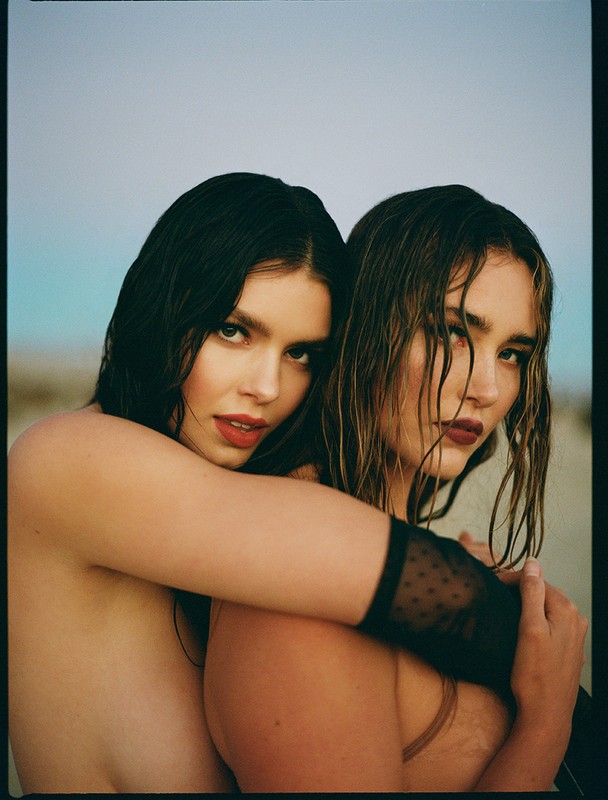 Two women embrace in an outdoor fashion shot. Brunette woman hugs a woman with highlighted hair against a neutral and blue background.