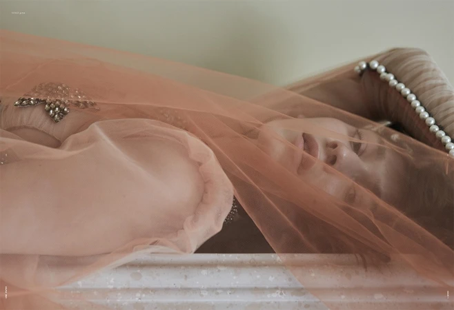 A woman lying down under a sheer peach-colored tulle veil with pearl embellishments on a headboard.