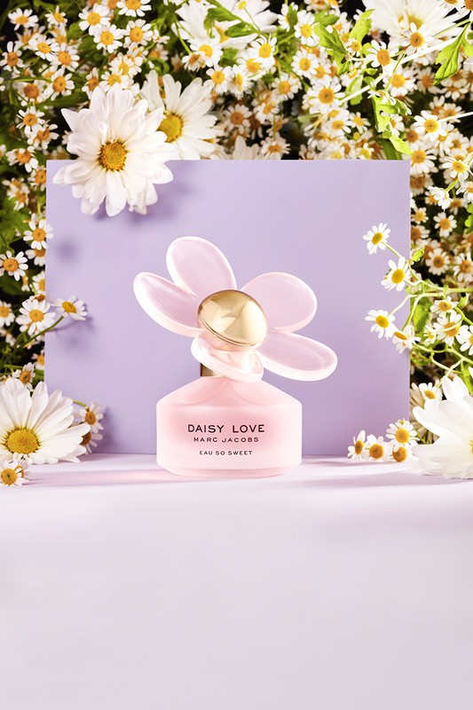 Close-up of a pink 'Daisy Love Marc Jacobs Eau So Sweet' perfume bottle with a daisy-shaped cap, against a floral background.