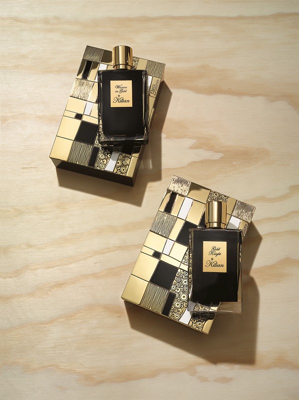 Two black Kilian perfume bottles rest on decorative gold and black boxes on a light wood surface in a studio setting.