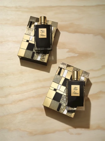 Two black Kilian perfume bottles rest on decorative gold and black boxes on a light wood surface in a studio setting.