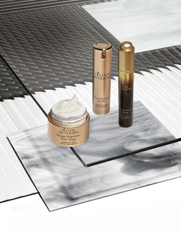 Three gold-toned skincare products, including a cream and two serums, are arranged on a geometric surface in a high-key beauty product shot.