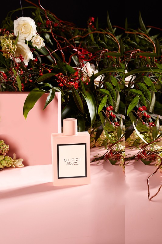 Gucci Bloom perfume bottle on a pink surface surrounded by white roses, green leaves, and red berries against a dark background.