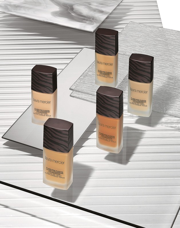Laura Mercier foundation bottles arranged on reflective surfaces and a white background with linear patterns, studio shot for beauty.