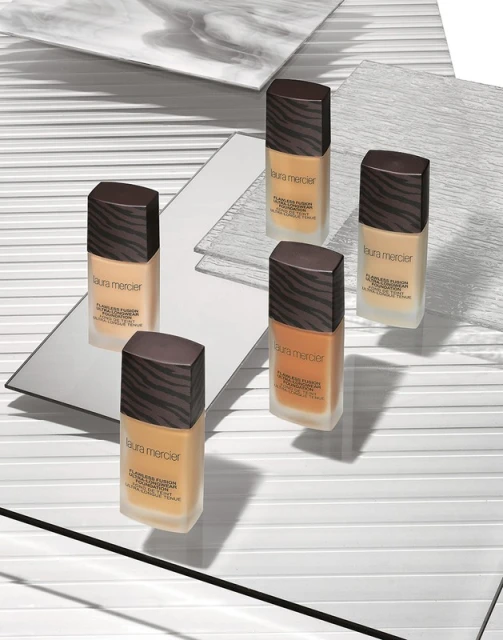 Laura Mercier foundation bottles arranged on reflective surfaces and a white background with linear patterns, studio shot for beauty.