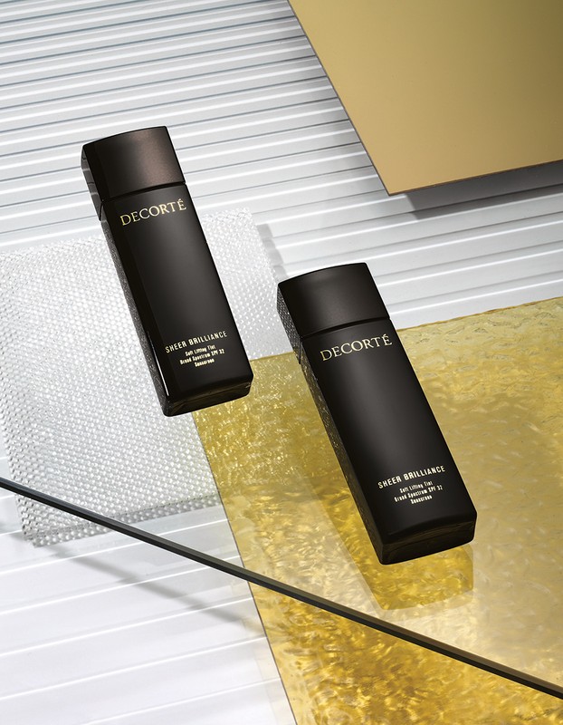 Two dark bottles of Decorté Sheer Brilliance foundation sit on a textured surface with gold and white accents in a beauty shot.