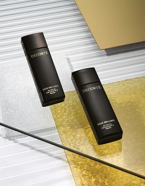 Two dark bottles of Decorté Sheer Brilliance foundation sit on a textured surface with gold and white accents in a beauty shot.