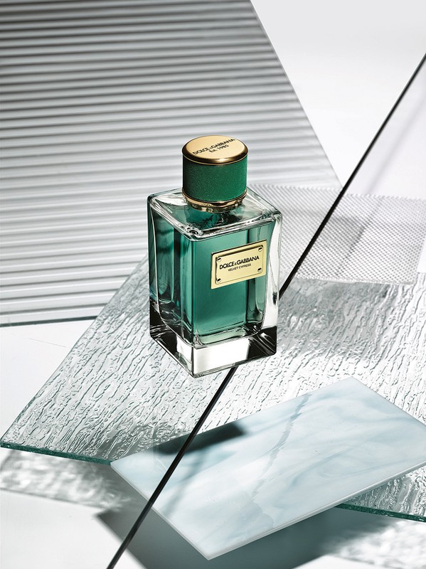 Green glass perfume bottle, gold cap, rests on clear textured plates, against striped white backdrop, in a studio-lit beauty product shot.