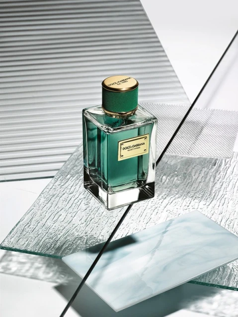 Green glass perfume bottle, gold cap, rests on clear textured plates, against striped white backdrop, in a studio-lit beauty product shot.