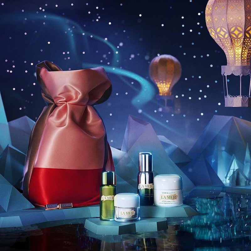 La Mer skincare products featured with a pink satin gift bag, set against a fantasy background with glowing hot air balloons.