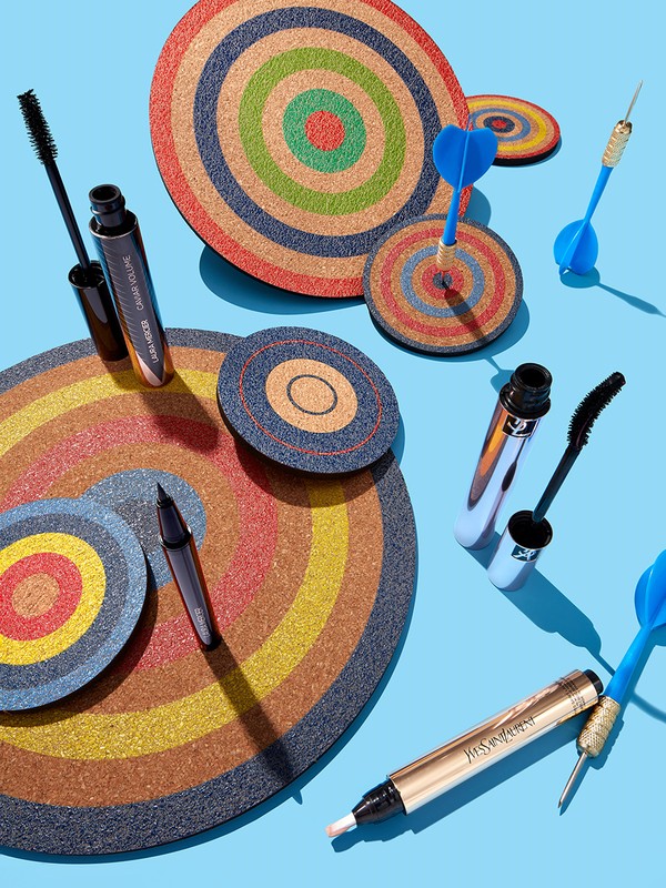 Beauty still life: cosmetic products including mascara and eyeliner arranged with colorful dartboard targets on bright blue backdrop.