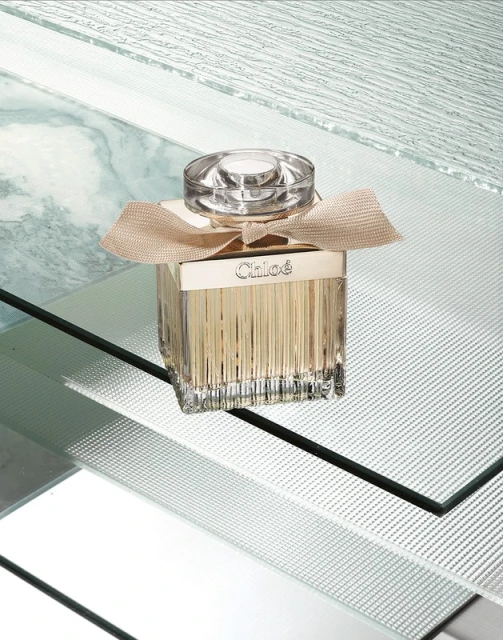 Chloe perfume bottle on reflective surface featuring its geometric glass design and tan ribbon detail.