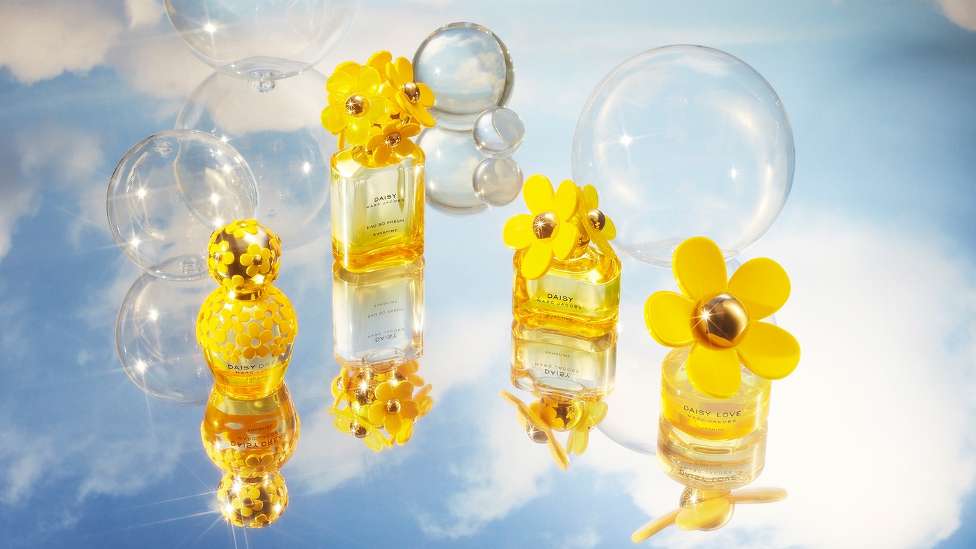 Close-up of four Daisy Marc Jacobs perfume bottles with yellow floral caps, set on a reflective surface against a sky backdrop.