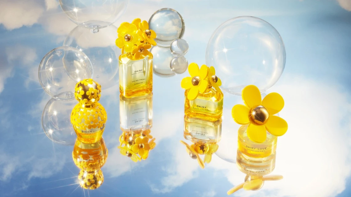 Close-up of four Daisy Marc Jacobs perfume bottles with yellow floral caps, set on a reflective surface against a sky backdrop.