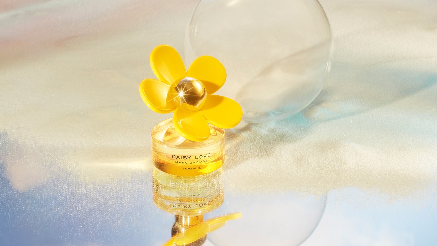 Bottle of Daisy Love Sunshine perfume by Marc Jacobs, with a yellow flower cap, on a reflective surface with a blurred background.