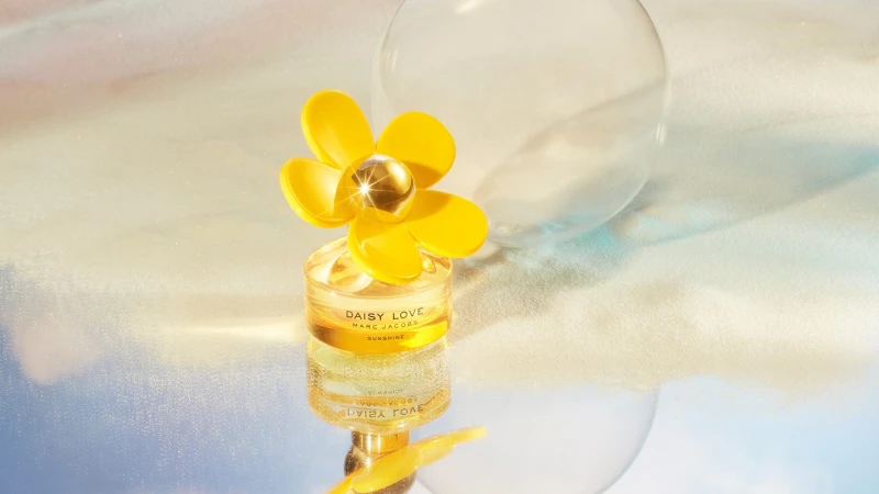 Bottle of Daisy Love Sunshine perfume by Marc Jacobs, with a yellow flower cap, on a reflective surface with a blurred background.