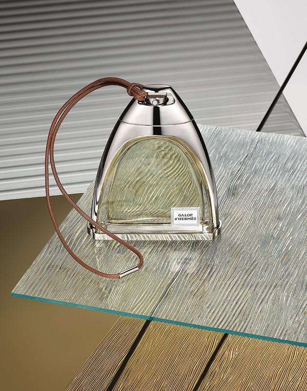 Elegant studio shot of a Galop d'Hermès perfume bottle with a silver cap and leather lanyard on a textured glass surface. 