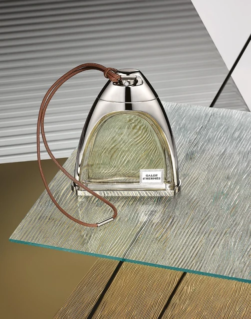 Elegant studio shot of a Galop d'Hermès perfume bottle with a silver cap and leather lanyard on a textured glass surface. 
