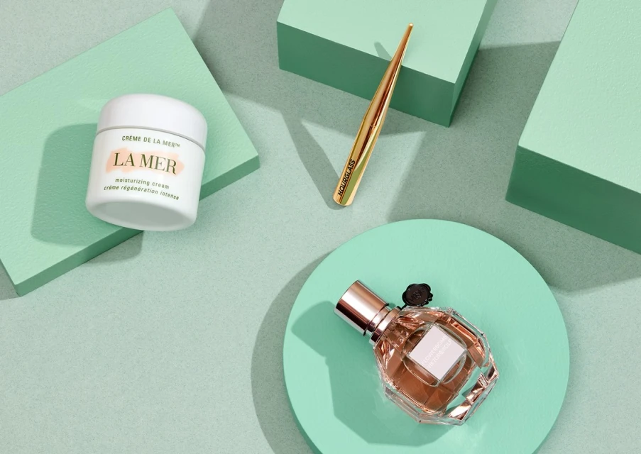 Flat lay of La Mer cream, Hourglass eyeliner, and Viktor & Rolf perfume on geometric mint-green platforms in a studio setting.