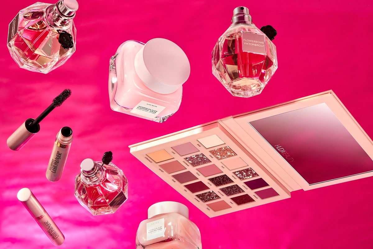 Cosmetics arranged on a pink background featuring perfume, eyeshadow, mascara, concealer, and moisturizer in a beauty product showcase.