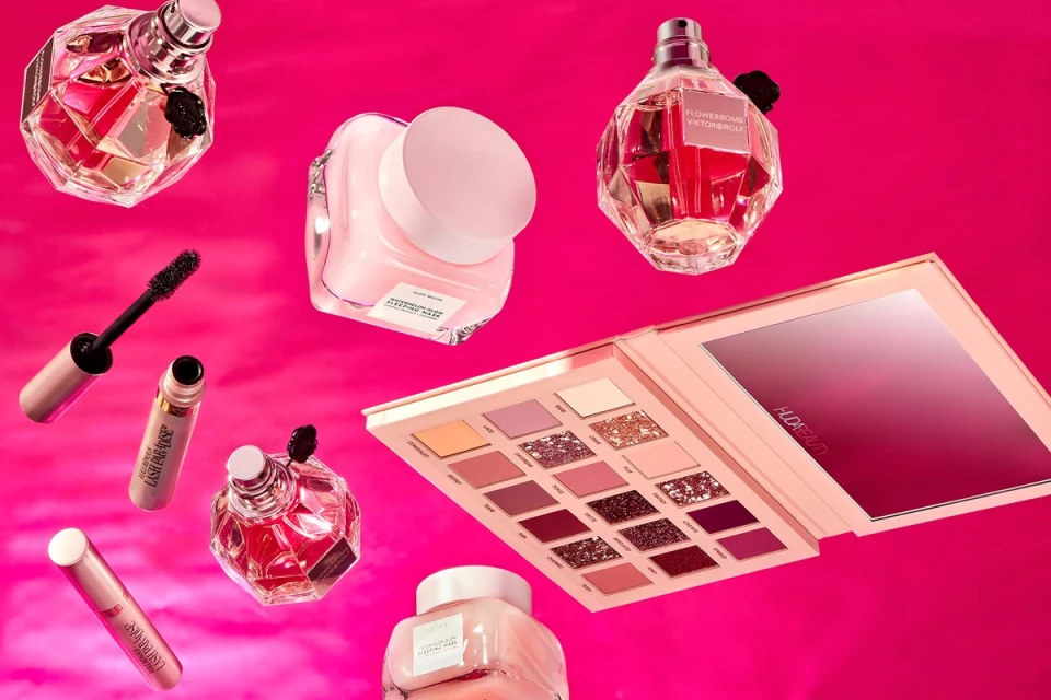 Cosmetics arranged on a pink background featuring perfume, eyeshadow, mascara, concealer, and moisturizer in a beauty product showcase.