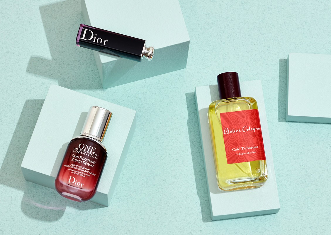 Dior lipstick, serum, and Atelier Cologne perfume bottles arranged on pale blue pedestals against a light blue background in a still life product shot.