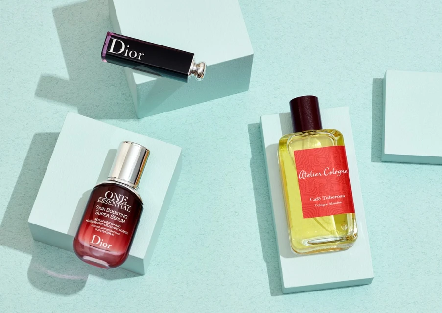 Dior lipstick, serum, and Atelier Cologne perfume bottles arranged on pale blue pedestals against a light blue background in a still life product shot.