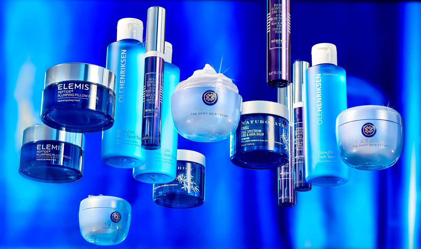 An array of blue-toned beauty products including creams, toners, and eye serums, arranged on a blue background. Brands are Elemis, Ole Henriksen, Tatcha.