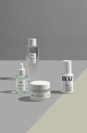 Still life shot of Huxley brand skincare products including facial oil, toner, and cream on a two-tone neutral surface against a grey background.