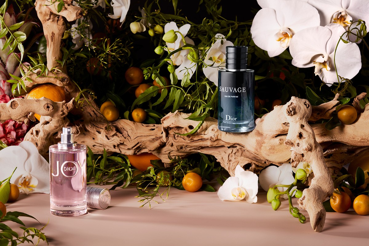 Two Dior perfume bottles, Joy and Sauvage, sit on a wooden branch surrounded by white orchids, greenery, and small oranges on a pink surface.