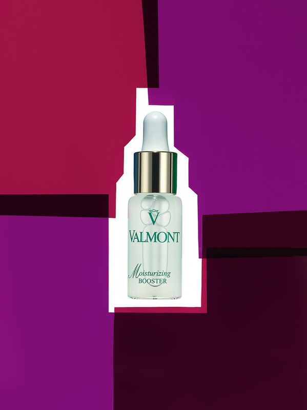 Close-up of Valmont Moisturizing Booster bottle with white and gold cap against a purple and red graphic background, skincare product.