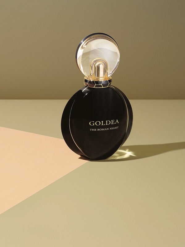 Round black perfume bottle with gold cap standing on a geometric beige and green studio surface with sharp shadows