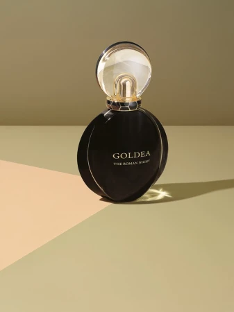 Round black perfume bottle with gold cap standing on a geometric beige and green studio surface with sharp shadows