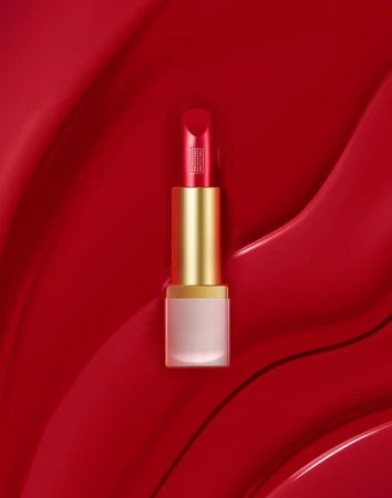 A red lipstick tube styled with prop styling by Alex Brannian set against a glossy red textured background.