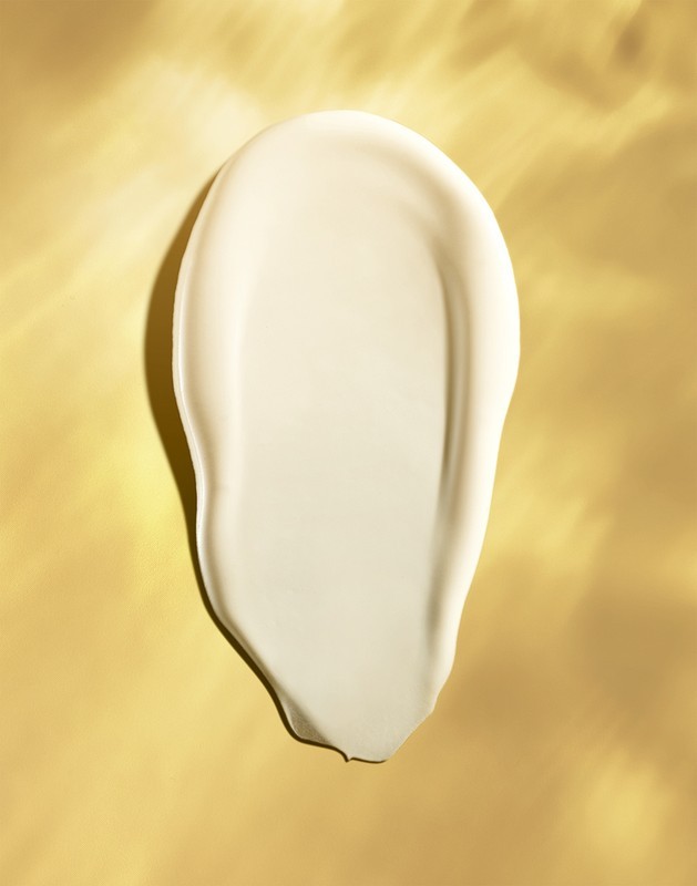 Close-up studio shot of a white cream swatch on a gold background. The swatch is smooth and shiny, catching light, in portrait orientation.
