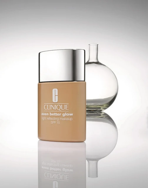 Clinique Even Better Glow foundation bottle in a studio product shot with a glass science flask, reflecting on a surface, and soft lighting.
