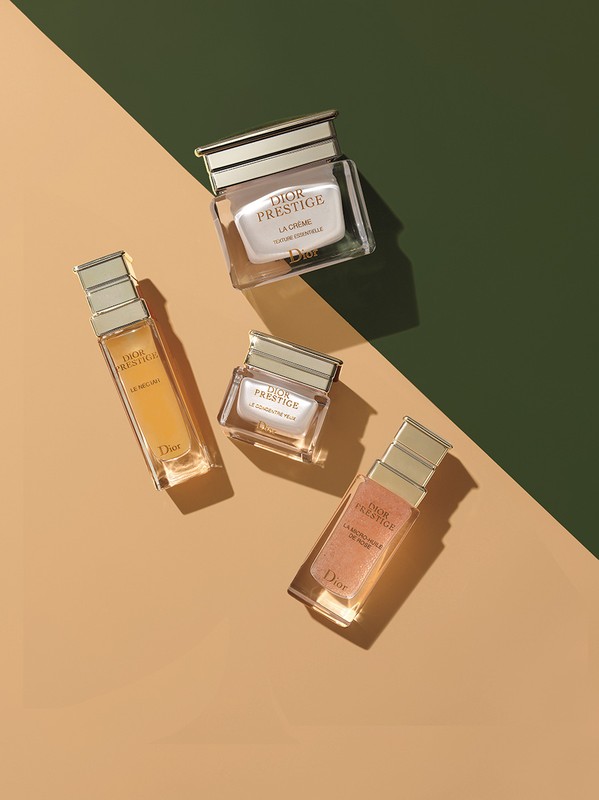 Four luxury skincare bottles and jars arranged diagonally on a two-tone tan and dark green geometric background.