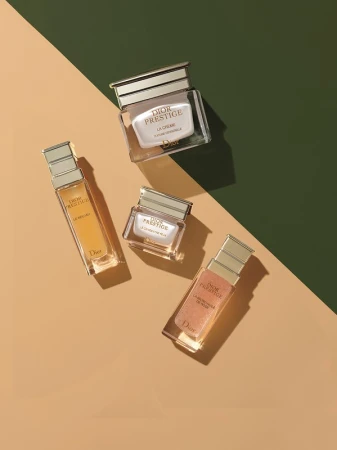 Four luxury skincare bottles and jars arranged diagonally on a two-tone tan and dark green geometric background.