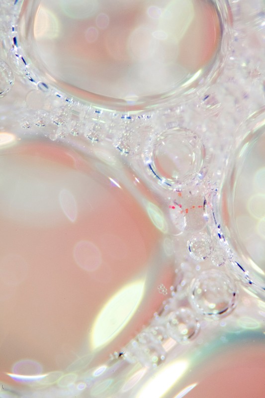 Macro photograph captures the intricate patterns of clear soap bubbles with iridescent highlights against a soft, pastel pink background.