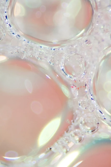 Macro photograph captures the intricate patterns of clear soap bubbles with iridescent highlights against a soft, pastel pink background.