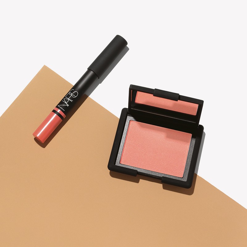 NARS makeup product set featuring blush compact and lipstick stick on a neutral tan and white background in a studio setting.