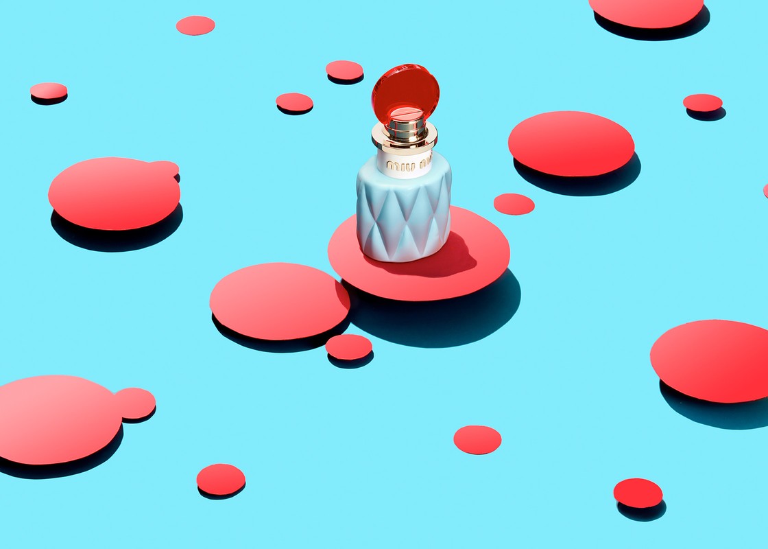 Studio shot of a light blue Miu Miu perfume bottle with a coral lid, presented on coral platforms, against a light blue background with coral dots.