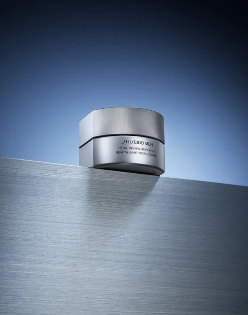 Shiseido Men Total Revitalizer Cream jar on a silver surface against a gradient blue backdrop, showcasing skincare product.