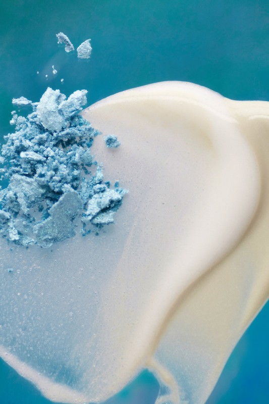 Close-up of light blue eye shadow powder alongside a stream of beige cream against a vibrant blue background.