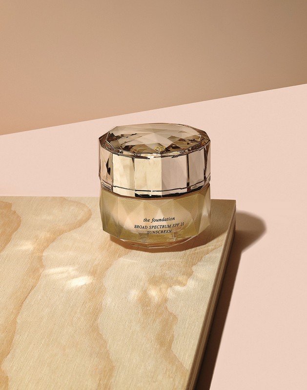 Close-up still life of skincare foundation in a jewel-faceted jar, placed on a light wood surface, against a tan backdrop.